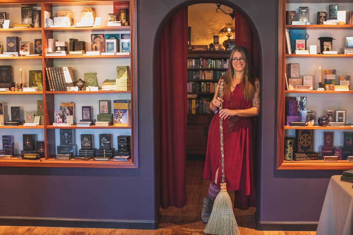 A woman in a red dress stands holding a broom in a cozy divination shop. Shelves filled with mystical books and items line the walls, evoking the metaphysical. A curtain behind her reveals more books and a warm, inviting atmosphere.