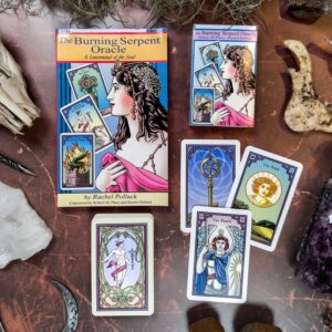 The image showcases The Burning Serpent Oracle with Book, beautifully arranged on a wooden surface, featuring captivating illustrations of a crowned woman, serpent, and burning chalice amidst enchanting crystals and feathers.