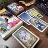 The Burning Serpent Oracle with Book set includes an oracle deck and box, featuring a card of a woman within a decorative border. Four cards depict the wheel, sun, and two figures. Crystals and stones are artfully placed around them.