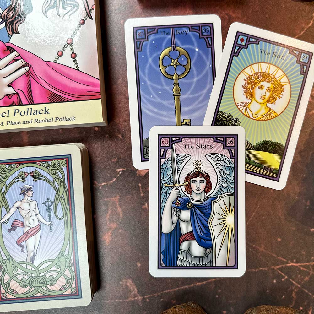 A oracle card spread featuring "The Key," "The Sun," and "The Stars" cards from The Burning Serpent Oracle, known for its intricate, colorful illustrations with symbolic imagery. A deck and book accompany the scene in the background.
