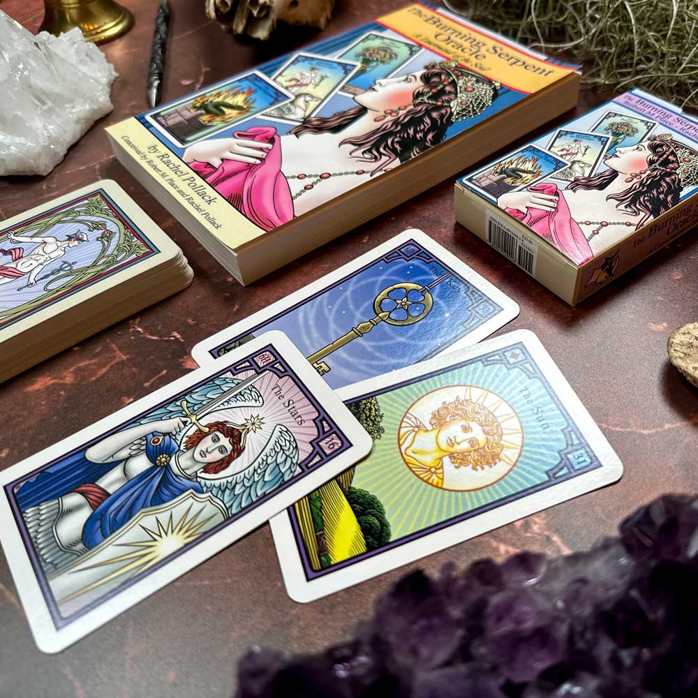 The Burning Serpent Oracle with Book lays on a marble table alongside a tarot card deck. Illustrated cards depict vivid scenes of a sun and winged figure, while crystals and mystical items complete the enchanting atmosphere.