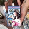 A person holds a box of "The Burning Serpent Oracle with Book," featuring an illustration of a crowned, bejeweled woman, while oracle cards are artfully arranged in the background on the table.