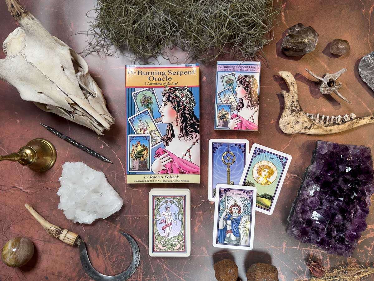 A mystical collection has oracle cards and two boxes of "The Burning Serpent Oracle with Book," featuring an illustrated figure. A skull, crystals, animal jawbone, candle holder, and sickle create an enchanting scene on the textured surface.