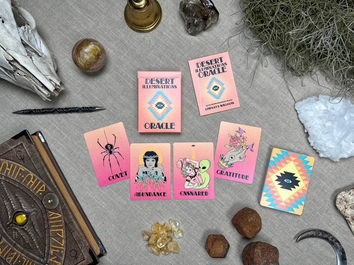 A flat lay of Desert Illuminations Oracle - 1st Edition tarot cards, crystals, a pen, feather, and candle on fabric. Cards show "Covet," "Abundance," "Ensnared," and "Gratitude" with illustrations. Natural elements and a patterned book surround them.