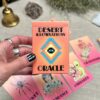 A person with black painted nails holds the vibrant "Desert Illuminations Oracle - 1st Edition" card pack. Cards depicting images and words such as "Coyote," "Abundance," and "Gratitude" are laid on a table, complemented by a bell and crystal.