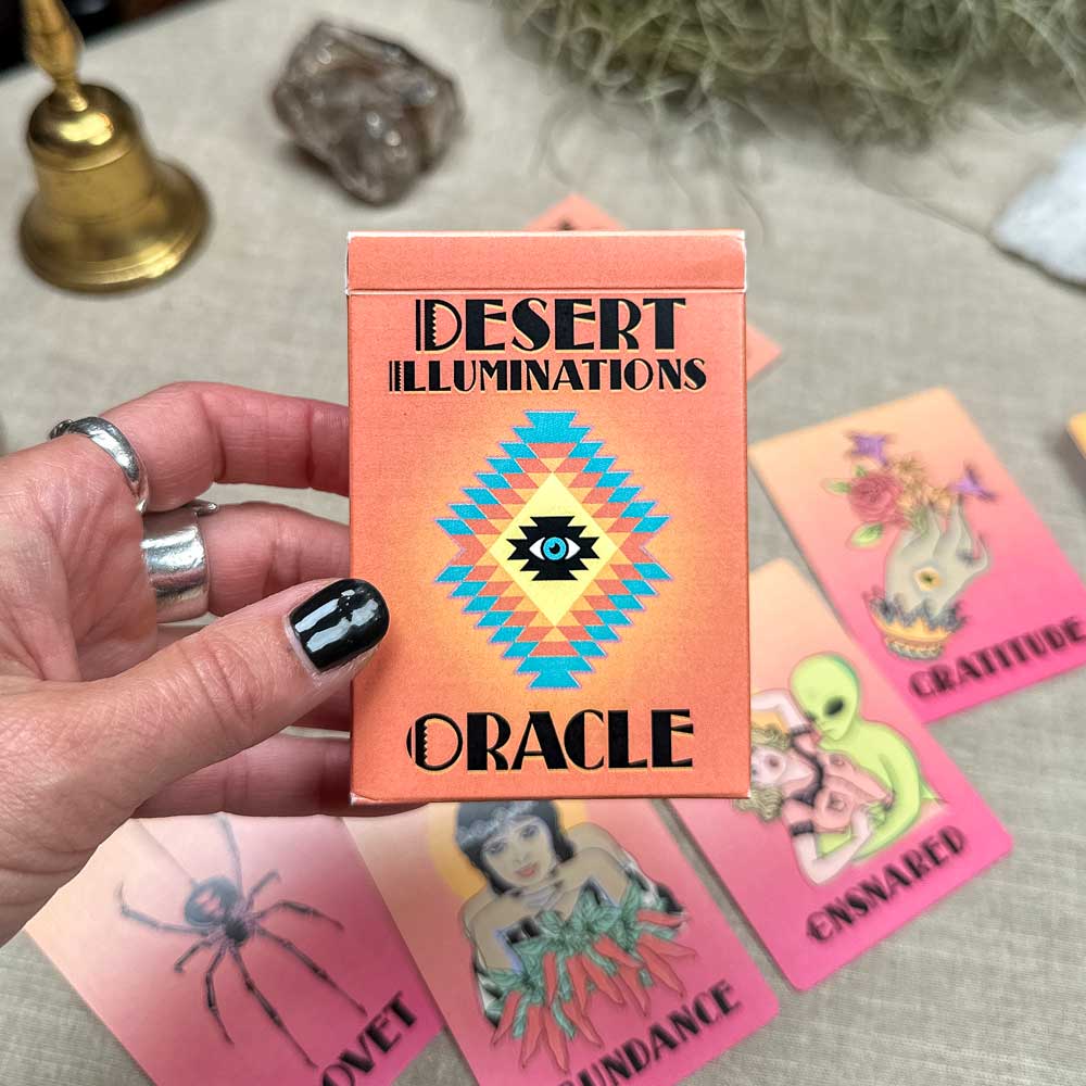 A person with black painted nails holds the vibrant "Desert Illuminations Oracle - 1st Edition" card pack. Cards depicting images and words such as "Coyote," "Abundance," and "Gratitude" are laid on a table, complemented by a bell and crystal.