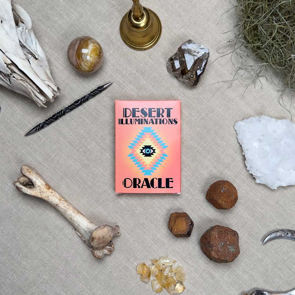 A flat lay artfully displays the “Desert Illuminations Oracle - 2nd Edition” amid bones, crystals, stones, a candle holder, a curved horn, and a decorative pen on a beige backdrop.