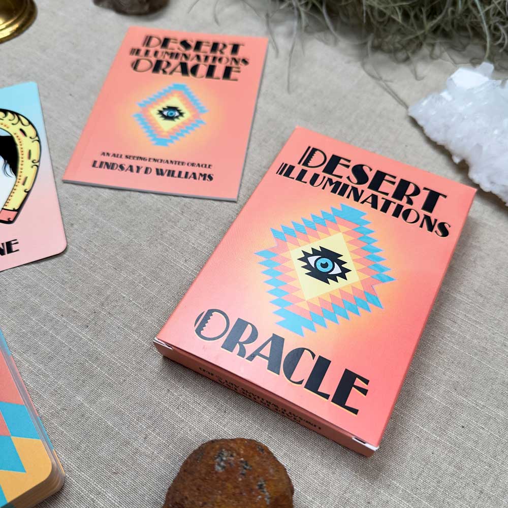 A "Desert Illuminations Oracle - 2nd Edition" box, featuring geometric designs and an eye illustration, sits on a table. A matching card is nearby amidst crystals and plants, evoking the mystical charm of the desert.