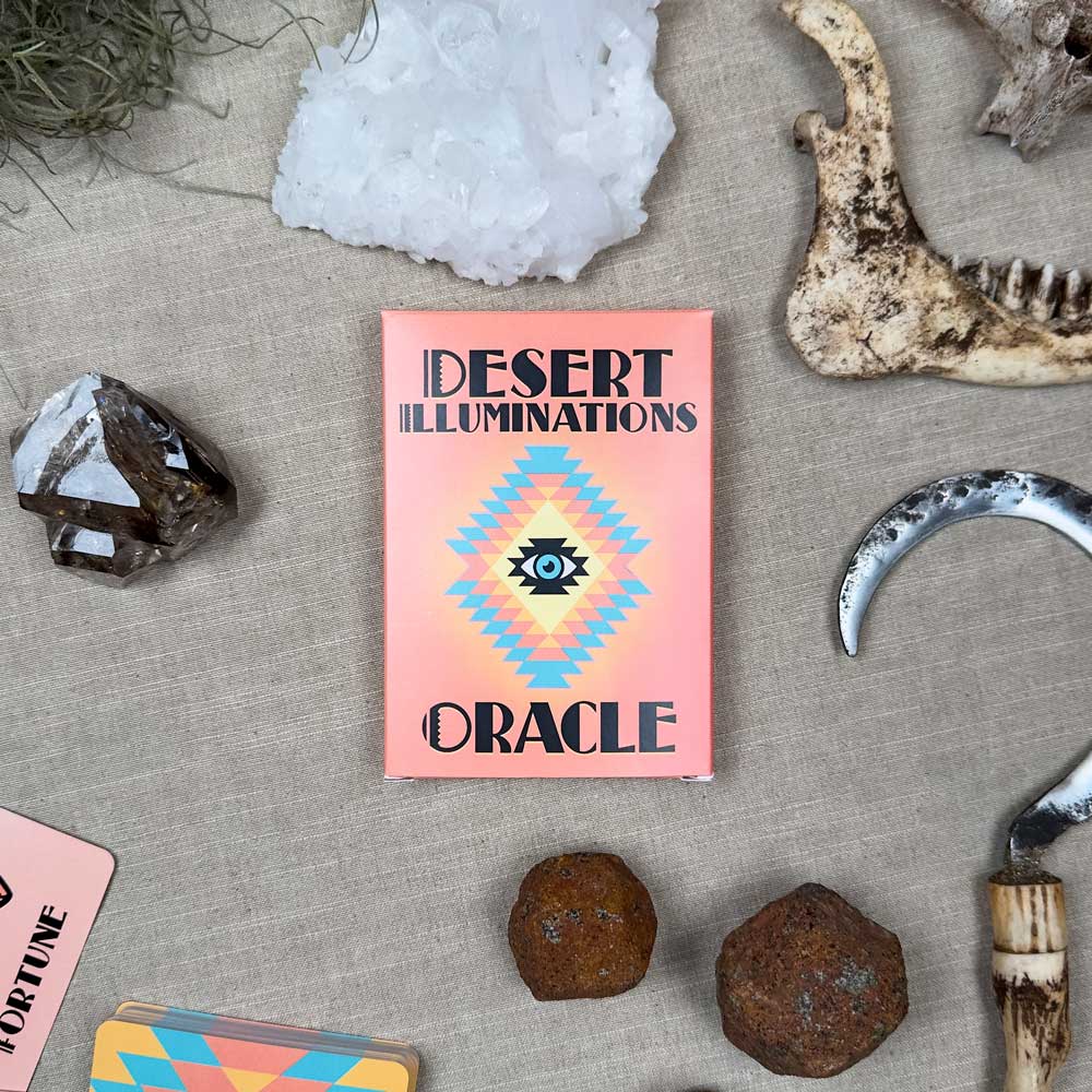 The "Desert Illuminations Oracle - 2nd Edition" box, adorned with a central eye in its geometric design, is surrounded by crystals, bones, a curved metal blade, and cards on a soft beige surface.