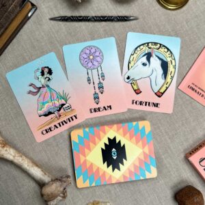 On a beige surface, three cards from the Desert Illuminations Oracle - 2nd Edition: "Creativity" featuring a woman, "Dream" with a dreamcatcher, and "Fortune" showcasing a horse head. The deck with geometric patterns is surrounded by a bone, pen, and candle.