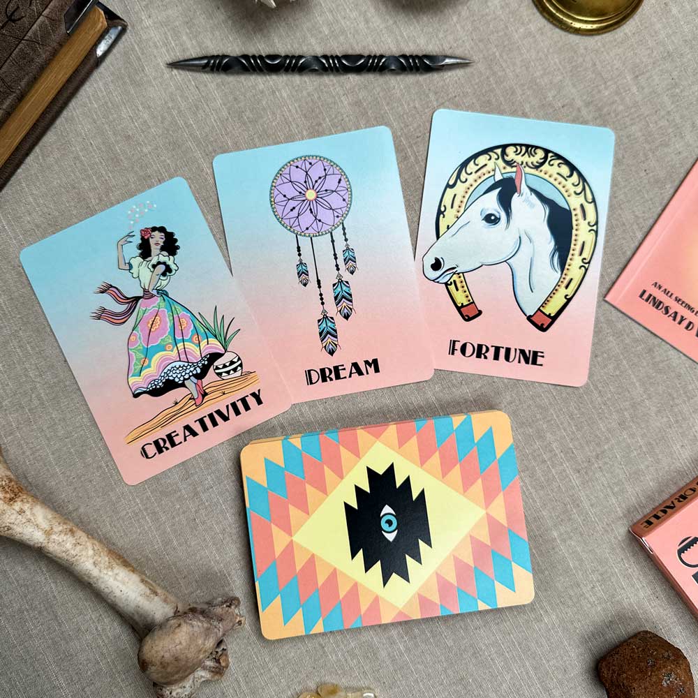 On a beige surface, three cards from the Desert Illuminations Oracle - 2nd Edition: "Creativity" featuring a woman, "Dream" with a dreamcatcher, and "Fortune" showcasing a horse head. The deck with geometric patterns is surrounded by a bone, pen, and candle.