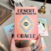 A hand adorned with rings holds a deck of Desert Illuminations Oracle - 2nd Edition cards. The box features a geometric design focused on an eye, while cards and books lie scattered across the table, ready for exploration.