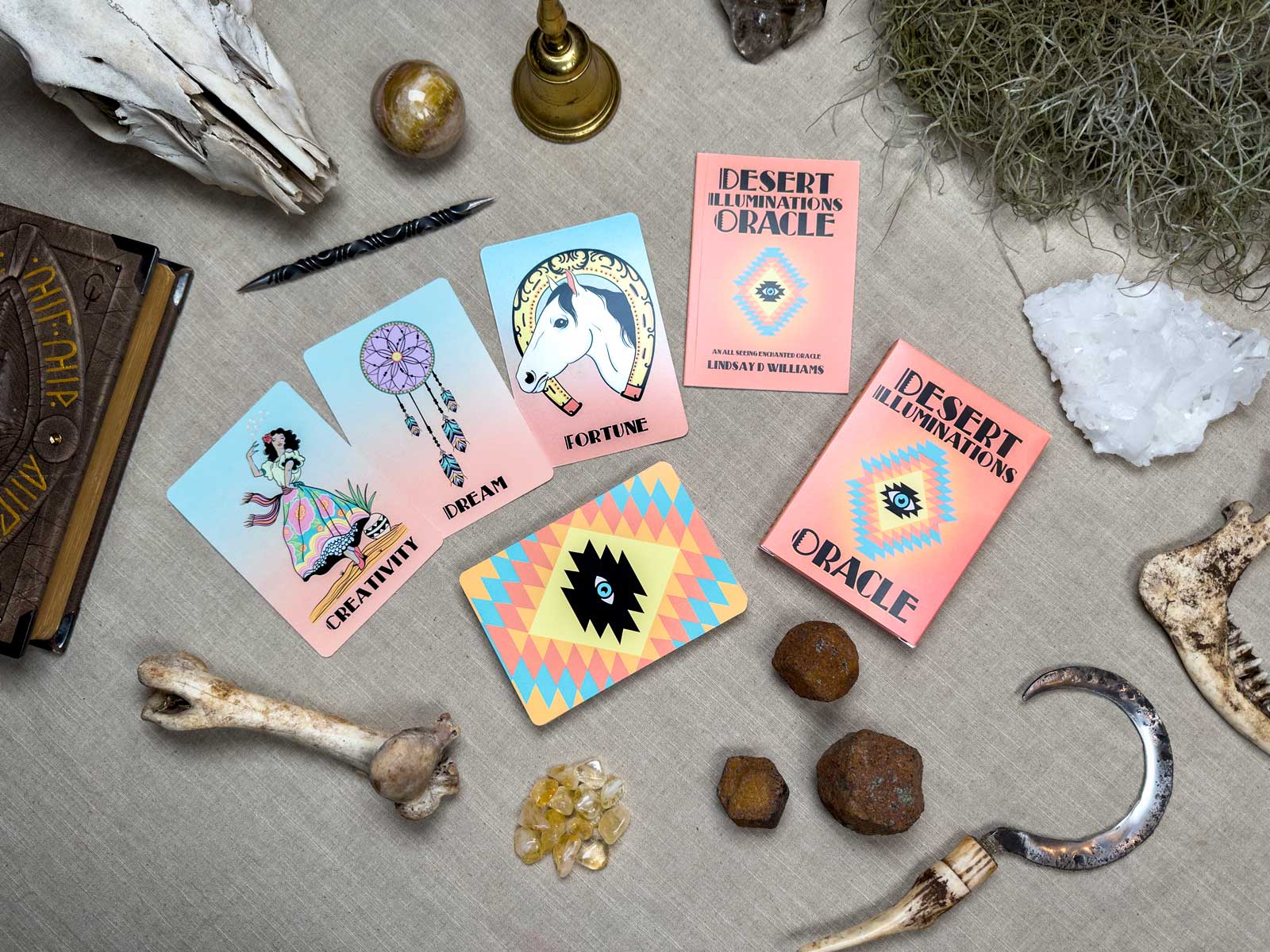 The table features Desert Illuminations Oracle - 2nd Edition cards with themes like "CREATIVITY," "DREAM," and "FORTUNE," surrounded by bones, crystals, a pocket watch, a candle, and books, crafting an enchanting mystery tableau.