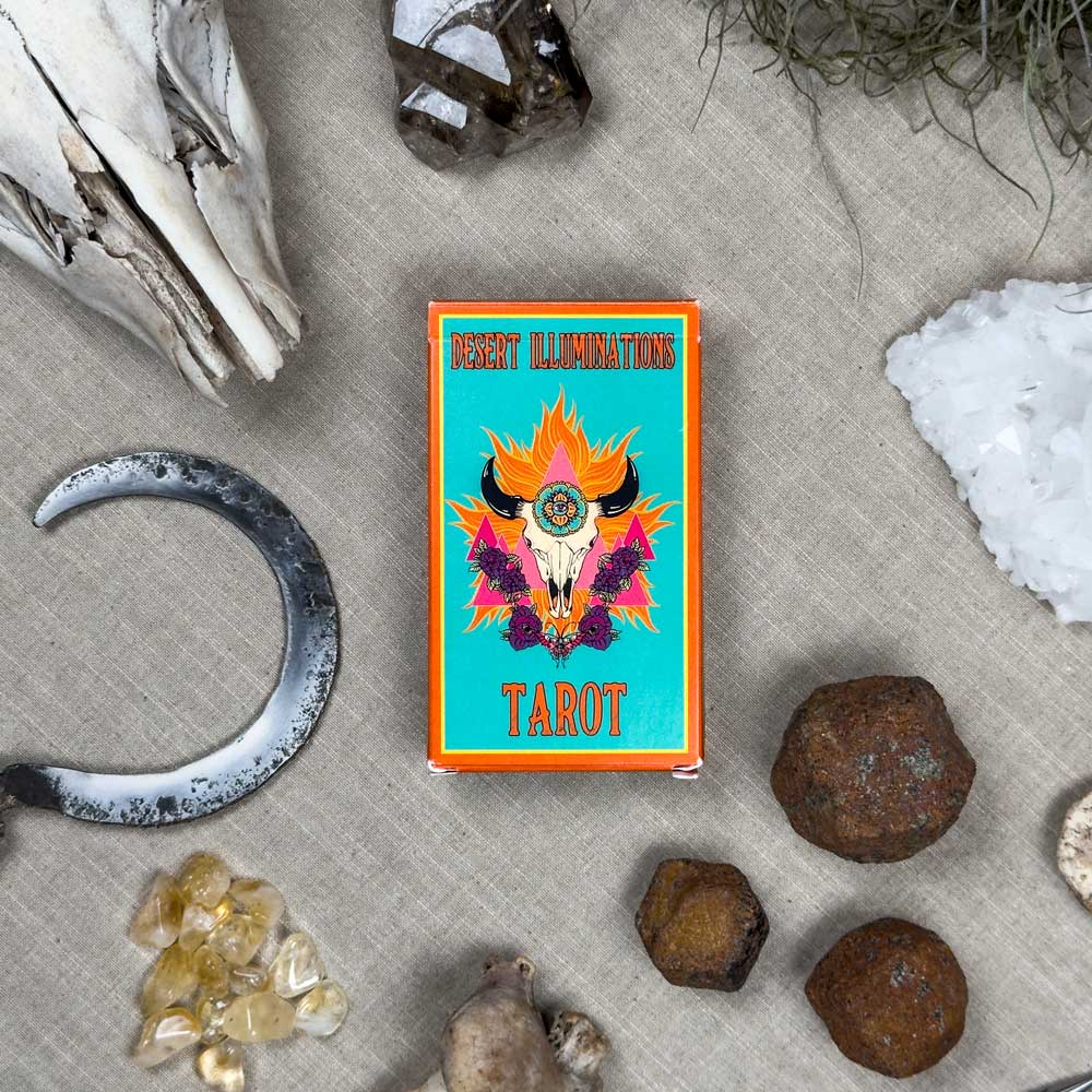 The "Desert Illuminations Tarot - 1st Edition" box rests on beige fabric, encircled by a crescent metal object, crystals, stones, a skull, and dried plants, creating a mystical ambiance.