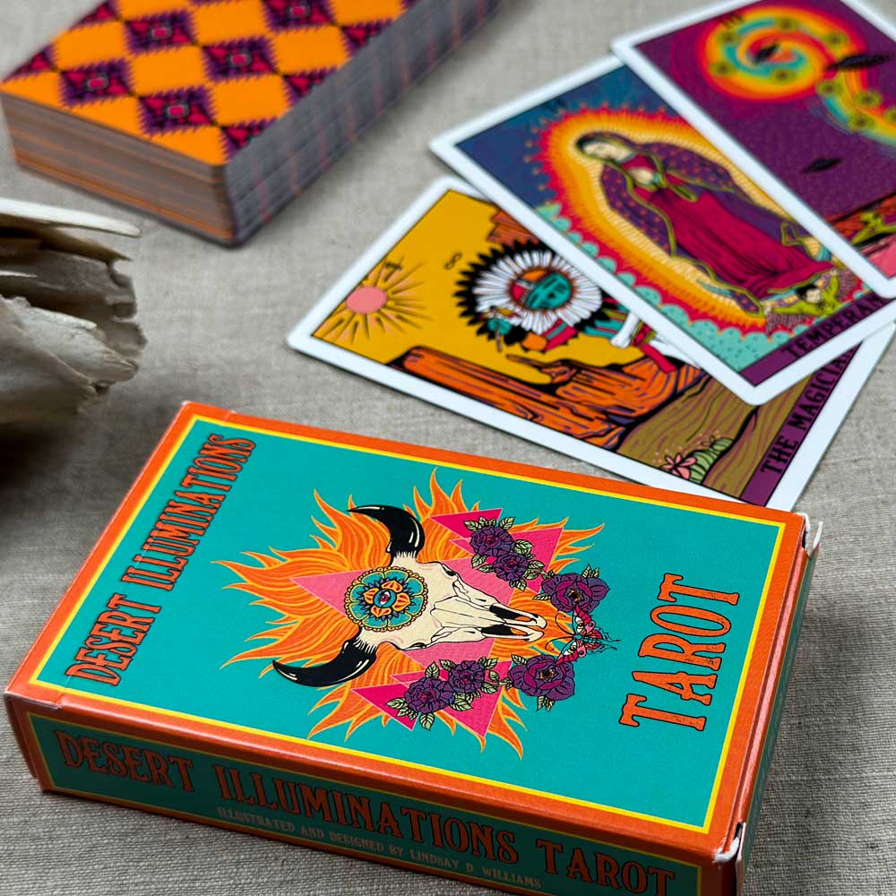 The "Desert Illuminations Tarot - 1st Edition," a vibrant deck showcasing bright designs and a cow skull on the box, lies on the table beside vividly illustrated cards and a crystal, enhancing the mystical atmosphere.
