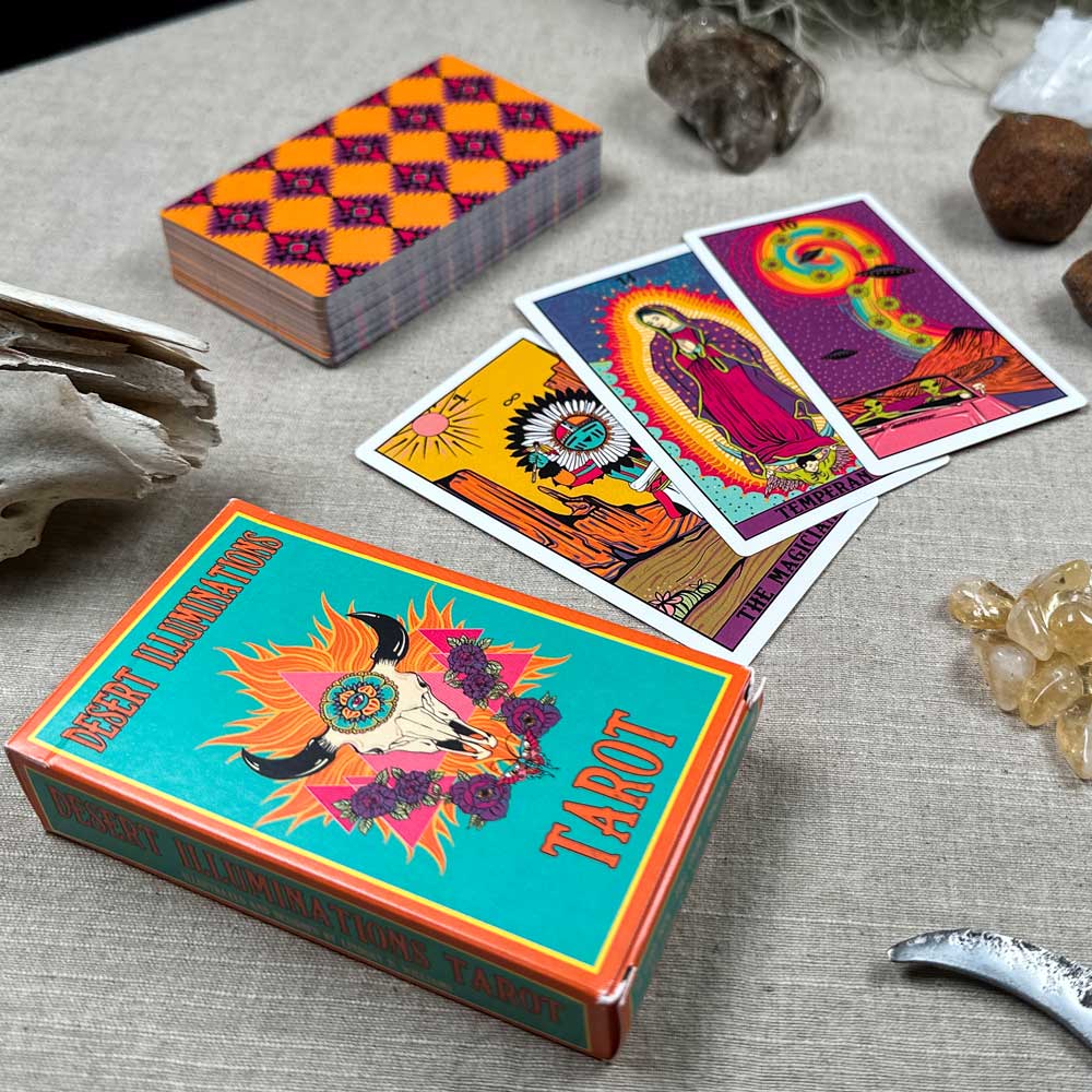 The Desert Illuminations Tarot - 1st Edition is displayed on a table. Three drawn cards showcase its colorful, mystical designs, while crystals and natural objects are artistically scattered around.