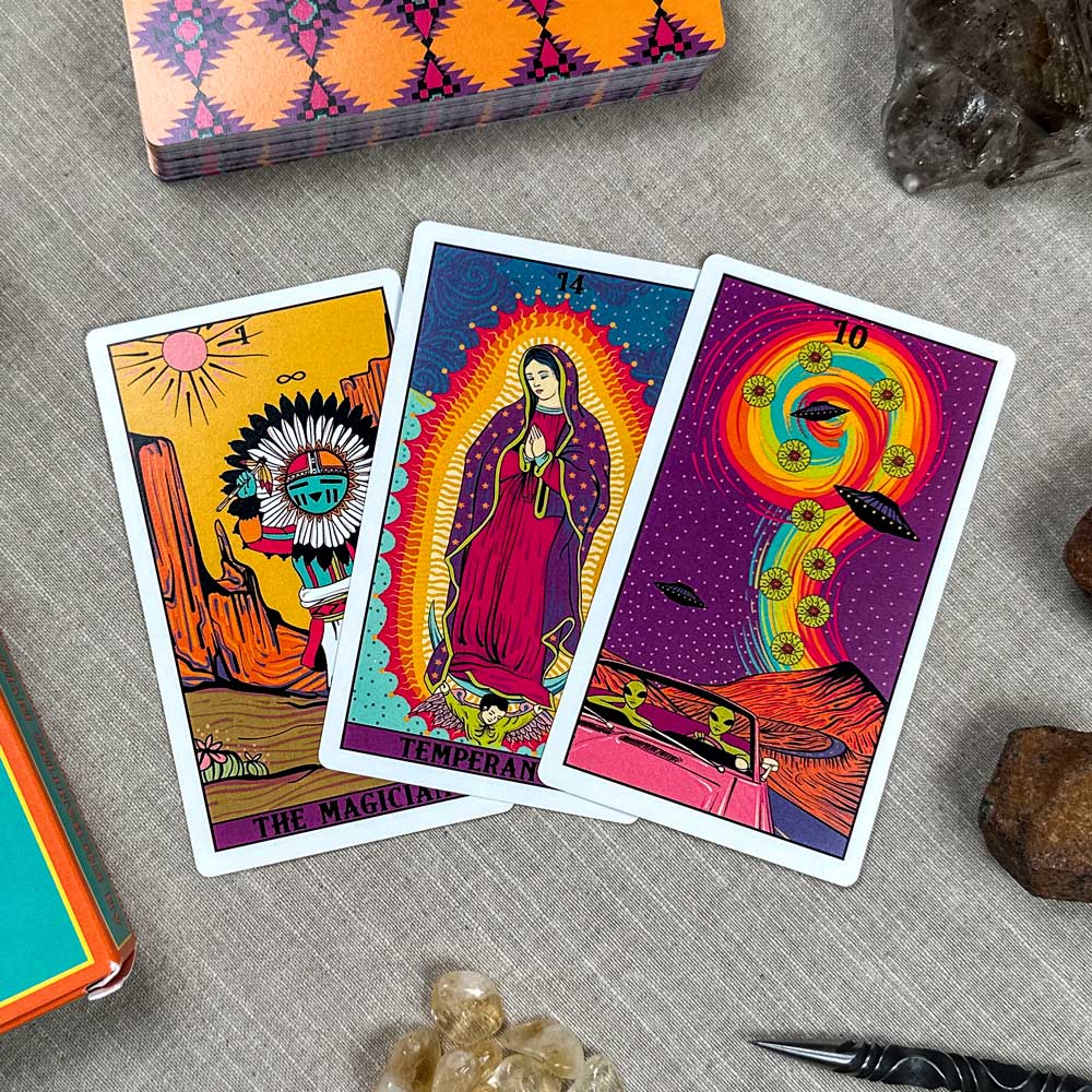 Three tarot cards—The Magician, Temperance, and Wheel of Fortune—featuring vibrant colors and cosmic themes are laid on a surface. They are surrounded by crystals, a pen, with the colorful Desert Illuminations Tarot - 1st Edition deck box in the background.