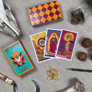 A flat lay of the Desert Illuminations Tarot - 1st Edition, showing The Magician, The High Priestess, and Ten of Wands cards, alongside a decorative box, crystals, bones, and stones on a textured surface.