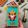Holding the "Desert Illuminations Tarot - 1st Edition" deck, a person displays its vibrant box adorned with a bull skull, flowers, and flames. Tarot cards, stones, and a bone enhance the mystical ambiance in the background.