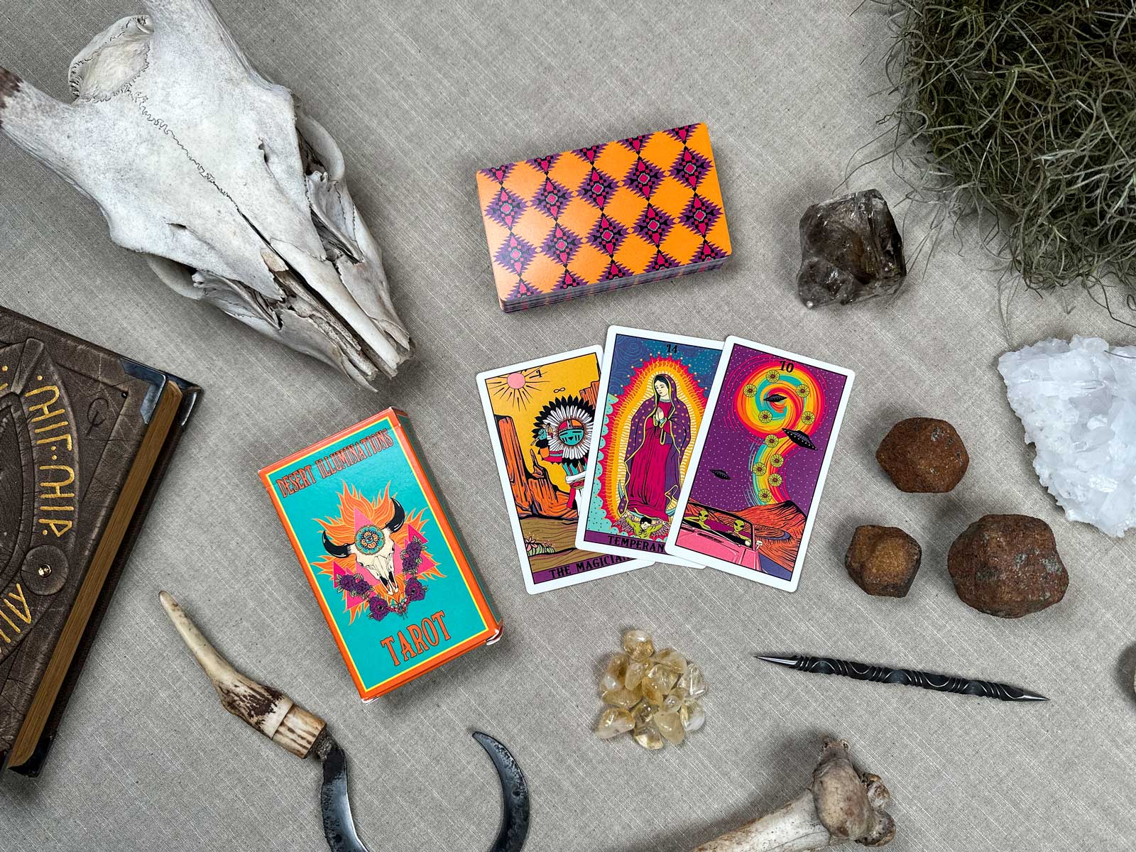 The Desert Illuminations Tarot - 1st Edition rests on a gray surface, showcasing three vibrant cards. Surrounding it are a patterned box, deer skull, crystals, rocks, a horn, knife, and an old book, creating a mystical and eclectic atmosphere.