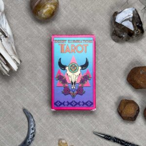 The "Desert Illuminations Tarot - 2nd Edition" box with southwestern motifs is on a textured surface, accompanied by crystals, a crescent moon-shaped item, and a small knife with an ornate handle.