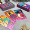 Tarot cards "The Trickster," "The Hierophant," and "Death" are spread on a table. Nearby is a colorful box labeled "Desert Illuminations Tarot - 2nd Edition," along with more cards and shimmering crystals in the ambient light.