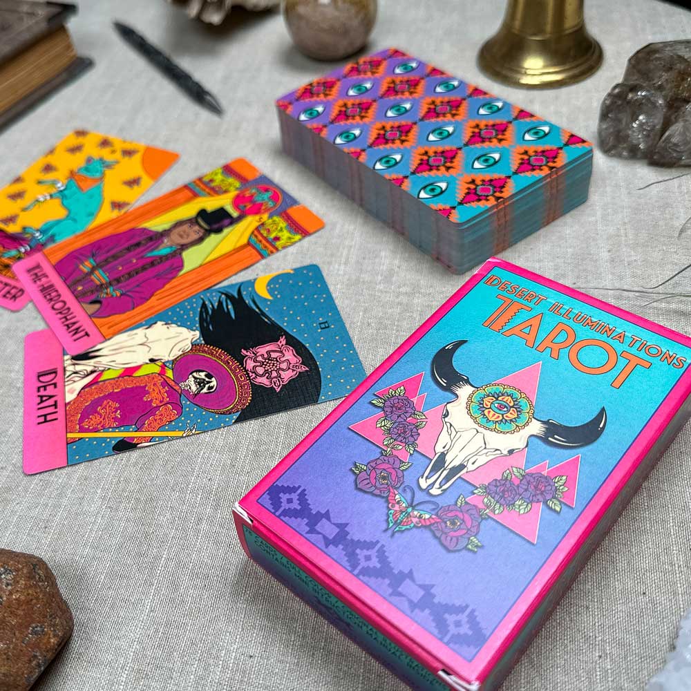 A close-up of the Desert Illuminations Tarot - 2nd Edition deck on a table shows colorful cards like "The Hermit" and "Death" fanned out, with the box featuring skull and flowers beside crystals and a pen on fabric.