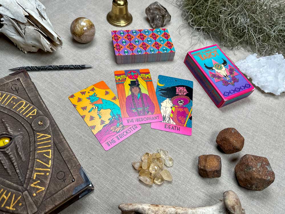 A table displays tarot cards such as "The Trickster," "The Hierophant," and "Death" from the Desert Illuminations Tarot - 2nd Edition. Nearby, a tarot deck box sits with a mystical symbol notebook, crystals, a brass candle holder, and natural items like rocks and moss on a beige cloth.