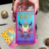 A hand holds the vibrant Desert Illuminations Tarot - 2nd Edition, featuring a box design with a flower-adorned cow skull. Background shows other tarot cards and mystical items partially visible on the table.