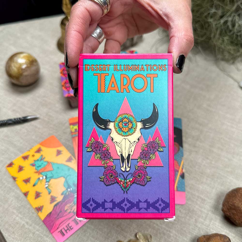 A hand holds the vibrant Desert Illuminations Tarot - 2nd Edition, featuring a box design with a flower-adorned cow skull. Background shows other tarot cards and mystical items partially visible on the table.