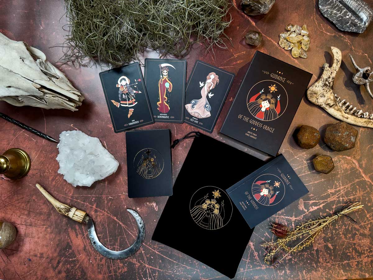 A collection of oracle cards, featuring the Guidance of the Goddess Oracle, is arranged on a textured surface. Nearby are a deer skull, assorted stones and crystals, a candle holder, a sickle, and dried herbs, creating an occult-themed setup.