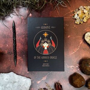 The "Guidance of the Goddess Oracle" box, set on a textured surface, is surrounded by shimmering crystals, a black wand, and dried plants for an enchanting mystical theme.