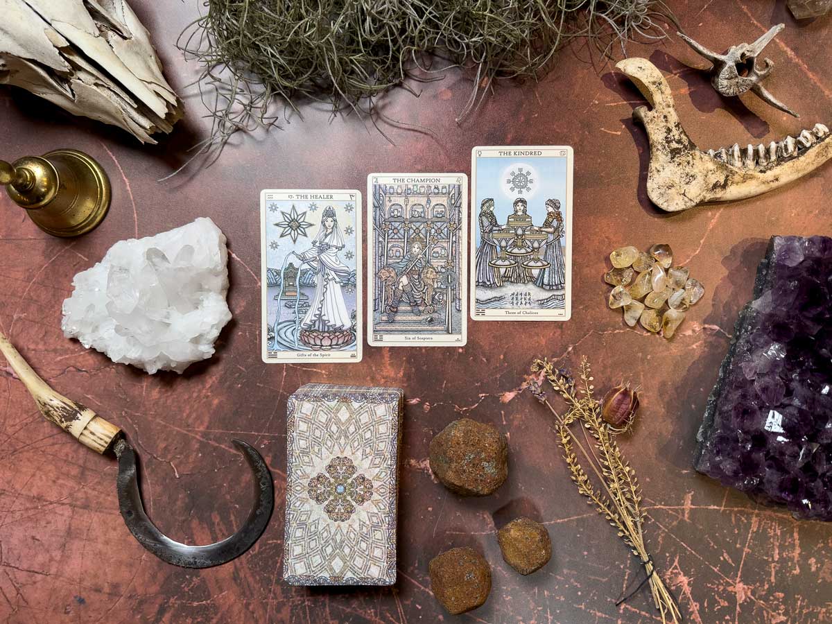 The Spirit Keepers Tarot Revelation Edition cards are spread on a table with crystal clusters, a brass candle holder, a sickle, dried plants, and bones. A patterned box complements the rustic setting, enhancing the mystical theme.