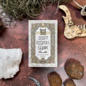 The "Spirit Keepers Tarot Revelation Edition" deck sits on a textured surface, encircled by crystals, bones, and rocks, highlighting its mystical aura in an earthy setting.
