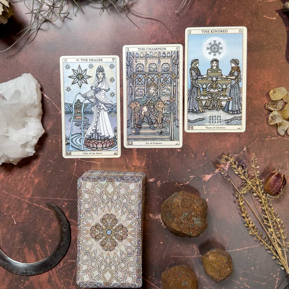 The Spirit Keepers Tarot Revelation Edition setup includes "The Healer," "The Champion," and "The Kindred" cards. Elegantly laid on a marbled surface, the display is surrounded by crystals, rocks, a decorative box, and a crescent-shaped object.