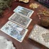 A display showcases three Spirit Keepers Tarot Revelation Edition cards on a textured surface, encircled by crystals, herbs, and a decorative tarot deck box. The cards feature intricate designs in muted colors, crafting a mystical and atmospheric ambiance.