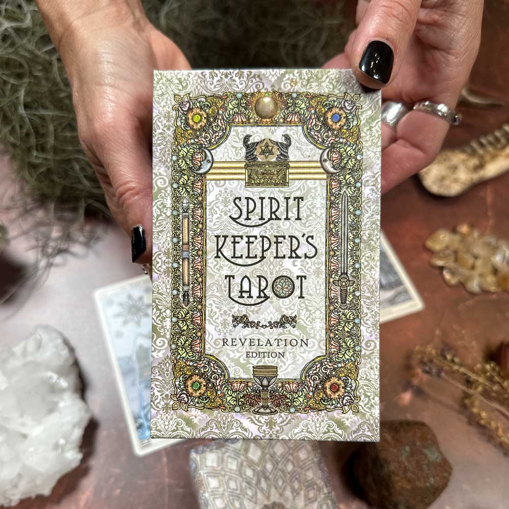 A person with black nail polish holds the "Spirit Keepers Tarot Revelation Edition." The ornate packaging features intricate designs, surrounded by crystals, gemstones, and decorative objects on a brown surface.