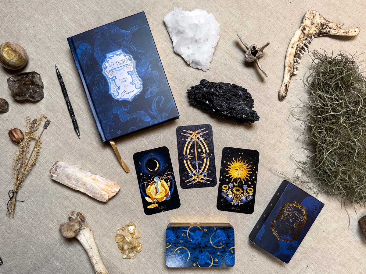 A mystical setup with Aurora Tarot cards, a journal, crystals, stones, and bones on beige fabric. The celestial and mythical designs of the Aurora Tarot cards paired with natural elements create an enigmatic ambiance.