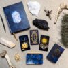 A mystical flat lay showcases the Aurora Tarot deck with celestial imagery, a blue book, crystals, bones, driftwood, all artistically laid out on beige fabric.