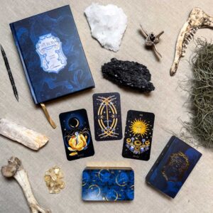 A mystical flat lay showcases the Aurora Tarot deck with celestial imagery, a blue book, crystals, bones, driftwood, all artistically laid out on beige fabric.