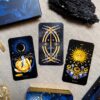 Three Aurora Tarot cards lie on a beige surface, joined by a sparkling crystal cluster, black stone, and decorative box. The cards feature vivid artworks: one with a moon and flower, another with crossed swords, and the last depicting a sun surrounded by floral patterns.