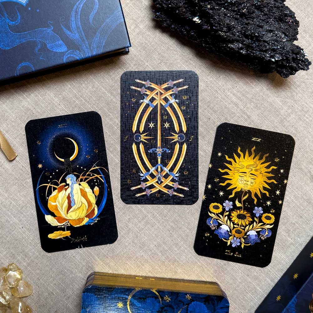 Three Aurora Tarot cards lie on a beige surface, joined by a sparkling crystal cluster, black stone, and decorative box. The cards feature vivid artworks: one with a moon and flower, another with crossed swords, and the last depicting a sun surrounded by floral patterns.
