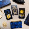 Four Aurora Tarot cards lie on linen, encircled by crystals. One card shows a crescent moon figure, another a floral sun, and the third a circular sword motif. A booklet and the box are nearby.
