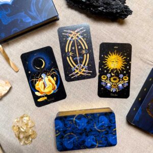 Four Aurora Tarot cards lie on linen, encircled by crystals. One card shows a crescent moon figure, another a floral sun, and the third a circular sword motif. A booklet and the box are nearby.