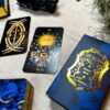 Displayed on a beige surface, a tarot setup features ornate cards, with one depicting "The Sun" adorned by intricate blue flowers. Nearby, the standout blue and gold Aurora Tarot box complements the natural surroundings.