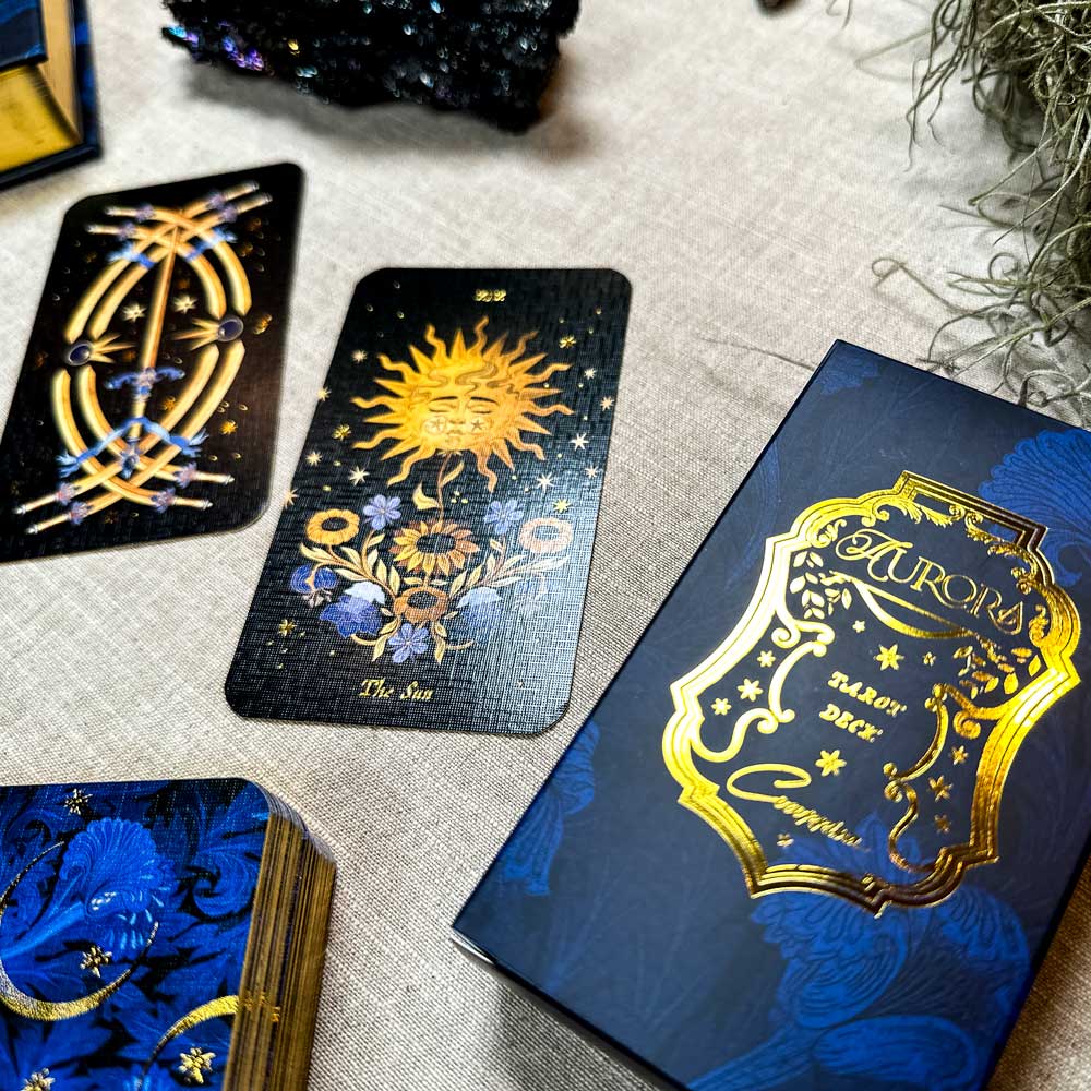 Displayed on a beige surface, a tarot setup features ornate cards, with one depicting "The Sun" adorned by intricate blue flowers. Nearby, the standout blue and gold Aurora Tarot box complements the natural surroundings.