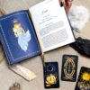 A book lies open with an illustration and text about Justice, marked by a gold ribbon. Beside it, three Aurora Tarot cards are displayed with crystals, a black rock, and a ring-adorned hand on a light fabric surface.