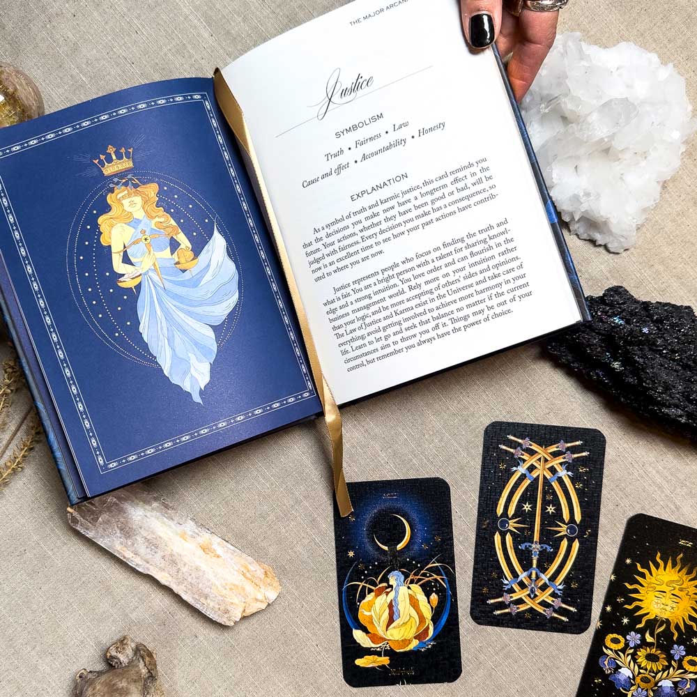 A book lies open with an illustration and text about Justice, marked by a gold ribbon. Beside it, three Aurora Tarot cards are displayed with crystals, a black rock, and a ring-adorned hand on a light fabric surface.