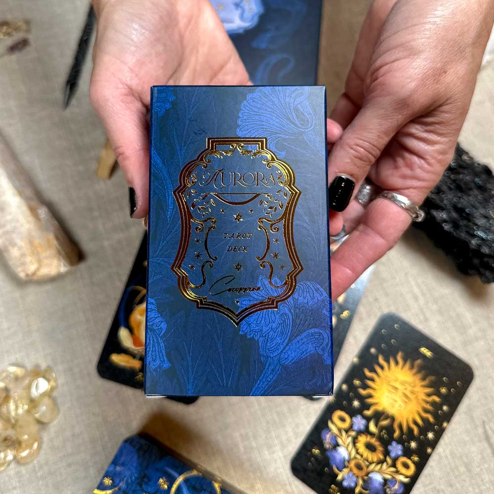 A person with black nail polish holds a blue and gold ornate "Aurora Tarot" box above a surface adorned with tarot cards, crystals, and stones.