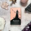 The Black Violet Tarot - Cherry Blossom Edition box displays a figure in a long black dress among birds and cherry blossoms, elegantly set on a surface with crystals and stones, exuding mystical allure.
